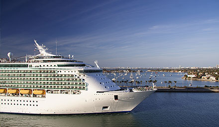 A generic cruise ship at a dock with tiny boats in the water with houses in a distance