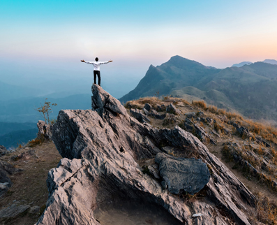Why Boscov’s Travel: a man on top of a mountain peak with arms outstretched under a clear blue sky