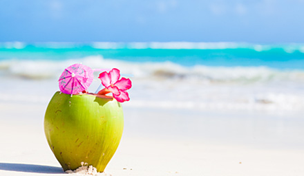 A green coconut drink with a pink drink umbrella and flower sitting on a sandy beach