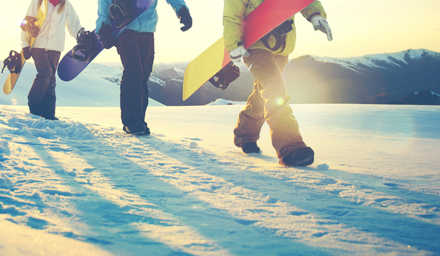 Three snowboarders carrying their snowboards in the snow with the sun setting in the background