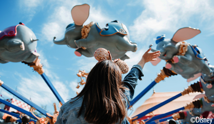A girl wearing sparkly rose gold Mickey ears waving at the Dumbo ride at Walt Disney World