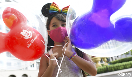 A girl wearing a colorful bow, wearing a mask, holding a red and purple Mickey ear balloon
