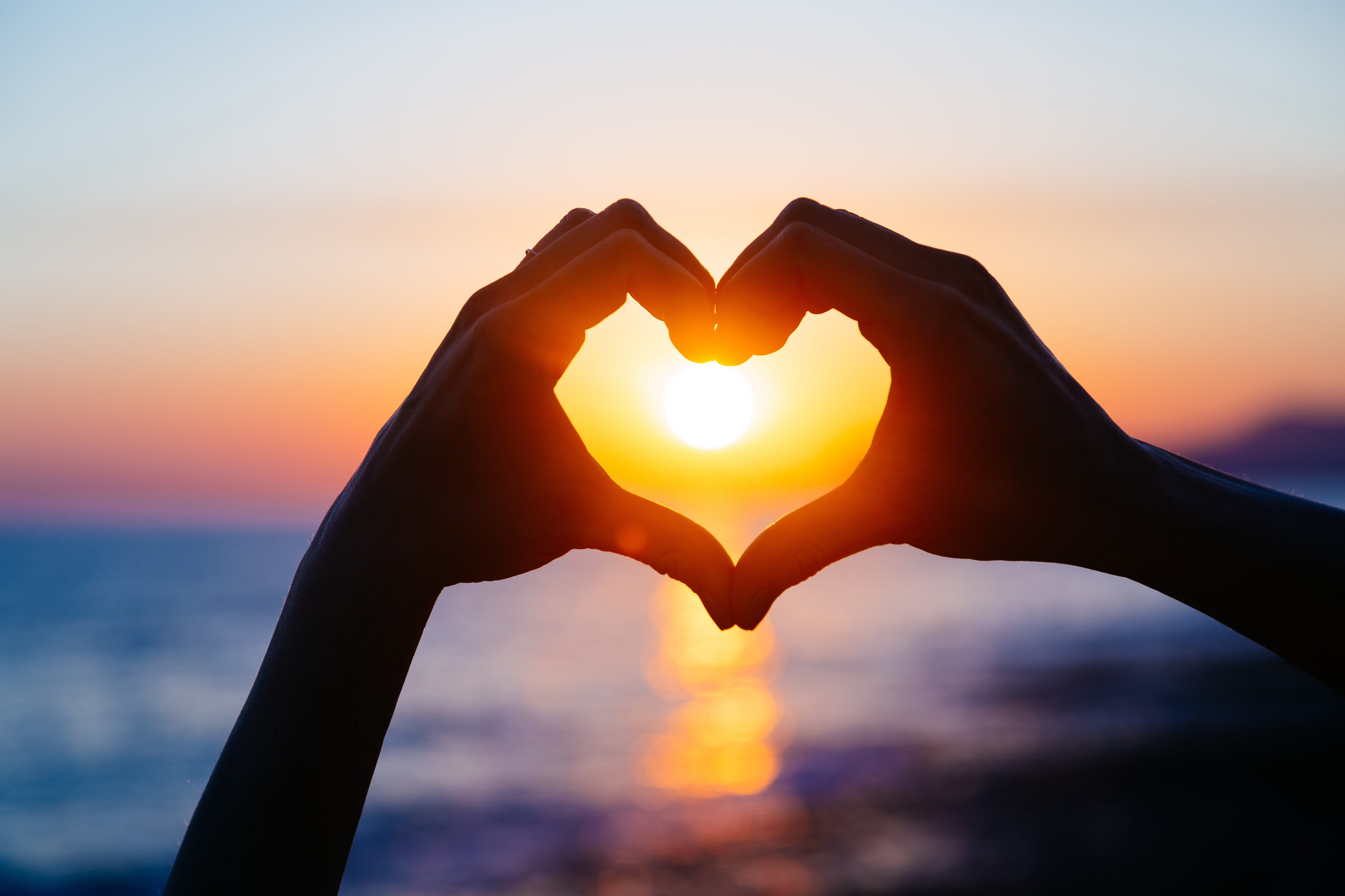 A silhouette of hands making the shape of a heart with the sun setting in the background