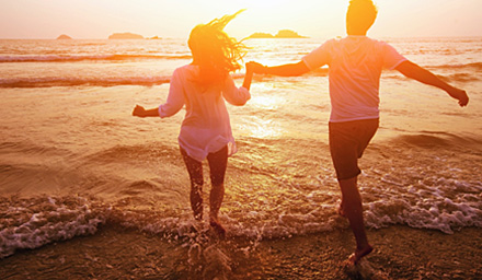 A cute couple holding hands running into the ocean with clothes on as the sun is setting in the background