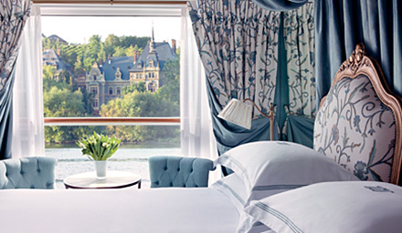 A bed with two blue chairs, table, and a window looking out to the water and a castle in the distance