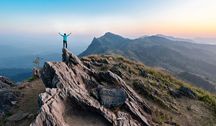 A man on top of a mountain peak with arms outstretched under a clear blue sky